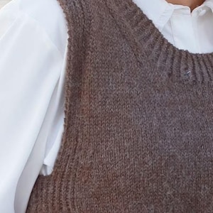 May include: A brown sleeveless knit vest featuring a ribbed crew neckline and armholes, worn over a crisp white collared shirt with long sleeves. The vest has a textured, soft-looking knit construction with a consistent dark brown colour throughout.