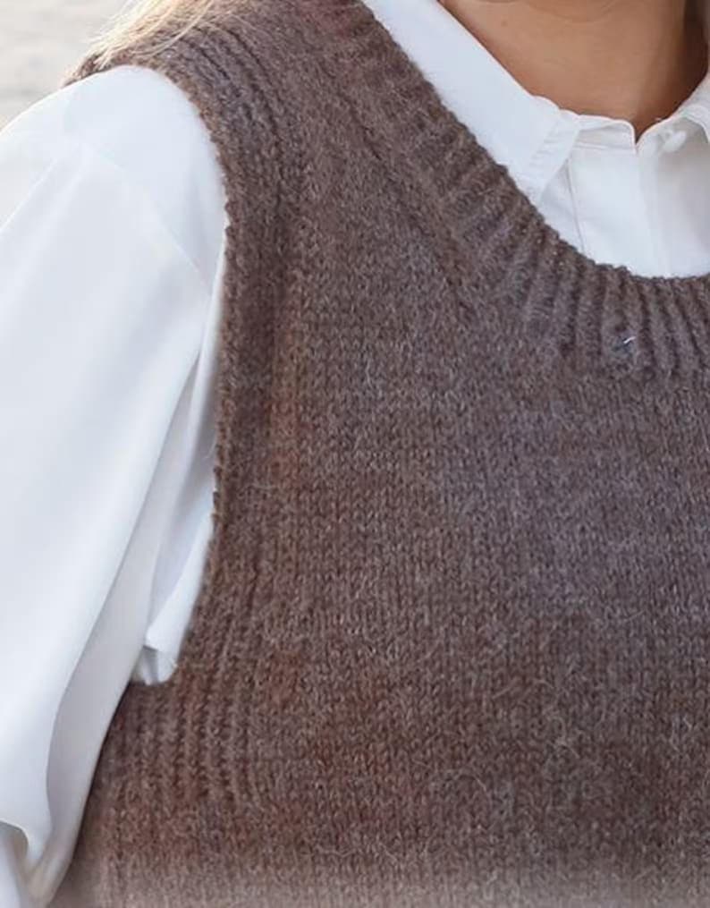 May include: A brown sleeveless knit vest featuring a ribbed crew neckline and armholes, worn over a crisp white collared shirt with long sleeves. The vest has a textured, soft-looking knit construction with a consistent dark brown colour throughout.
