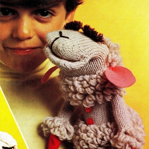 以下が含まれることがあります： ピンクの耳と赤い足が付いたグレーのニットの羊の人形。人形は茶色の髪の子供によって保持されています。