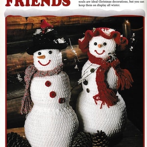 Op de afbeelding: Twee gehaakte sneeuwpoppen, een met een rode muts en sjaal, de andere met een zwarte muts en rode sjaal. Beide sneeuwpoppen hebben rode knopen als ogen en een wortelneus. De sneeuwpoppen zitten op een houten oppervlak met dennenappels.