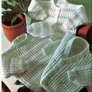 Op de afbeelding: Drie gebreide babyvesten in wit en mintgroen. De truien hebben knoopsluitingen en geribbelde details. De kledingstukken zijn gerangschikt op een groene achtergrond met een terracotta pot en plant zichtbaar.