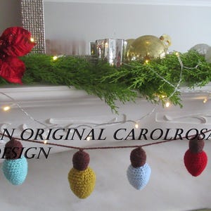 May include: A festive Christmas light garland featuring crocheted bulbs in red, blue, yellow, and green, strung on a brown cord. The garland is displayed on a white mantelpiece, decorated with greenery, a red poinsettia, and fairy lights. Text reads "AN ORIGINAL CAROLROSA DESIGN."