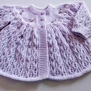 May include: A lilac-coloured, hand-knitted baby cardigan with long sleeves. The sweater has a textured openwork design, a round neckline, and small decorative embellishments near the collar. It has a button-up front and a flared skirt.