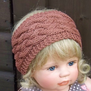 Puede incluir: Una diadema de punto de color rosa polvoriento con un patrón de punto trenzado. La diadema la lleva una muñeca con el pelo rubio y los ojos azules.