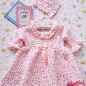 May include: A pink crocheted baby dress with a matching hat. The dress has a ruffled neckline and a bow at the waist. The hat is a simple beanie style.