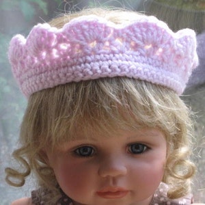 May include: A pink crocheted crown with a scalloped edge. The crown is worn by a doll with blonde hair and blue eyes.