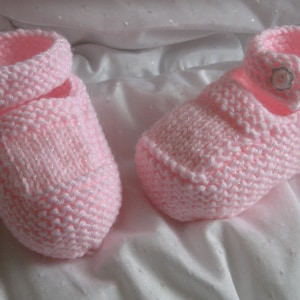 Pode incluir: Um par de sapatinhos de bebé de malha rosa com botões brancos. Os sapatinhos têm um design simples e são perfeitos para um recém-nascido.