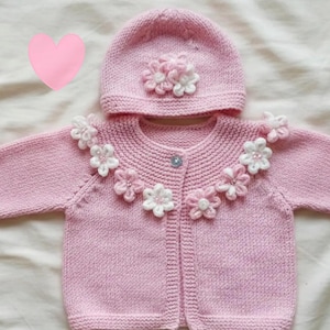Puede incluir: Conjunto de cárdigan y gorro de bebé de punto rosa claro. El cárdigan y el gorro están adornados con aplicaciones de flores blancas y rosas. El cárdigan tiene un cierre de un solo botón. El conjunto es para un bebé.