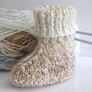 May include: A knitted baby bootie with a white cuff and a brown and white yarn pattern. The bootie is on a white surface with a ball of yarn in the background.