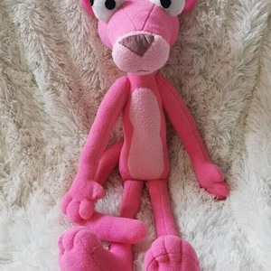 Può includere: Un peluche rosa a forma di pantera rosa, con grandi occhi espressivi, una pancia rosa chiaro e un naso marrone. Il giocattolo ha braccia e gambe lunghe ed è realizzato in materiale morbido e soffice. La pantera è un personaggio dei cartoni animati popolare.