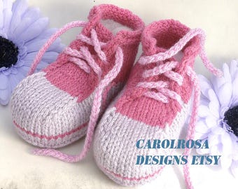 May include: A pair of hand-knitted baby booties in pink and light gray, featuring a lace-up design with pink yarn laces. The booties have a rounded toe and a ribbed cuff. The text CAROLROSA DESIGNS ETSY is printed in blue on the right side.