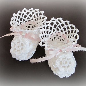 Puede incluir: Un par de botitas de bebé de ganchillo blanco con detalles de cinta rosa y adornos de perlas.