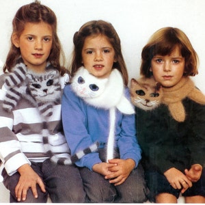 Könnte beinhalten: Drei Mädchen tragen gestrickte Kattenschals. Die Schals sind weiß, grau und braun mit Katzenköpfen. Die Mädchen tragen Pullover und Jeans.