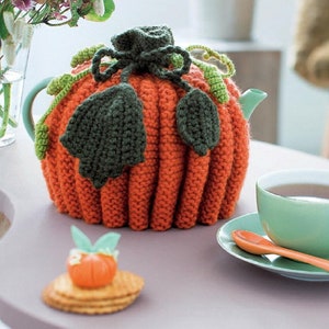 May include: A knitted orange pumpkin teapot cozy with green leaves. The cozy is on a table with a cup of tea and a small pumpkin-shaped cookie.