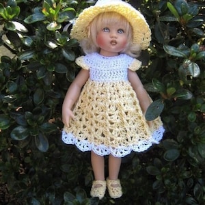 May include: A doll wearing a yellow and white crocheted dress with a white crocheted hat. The doll is standing in front of a green bush.