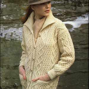 Peut inclure: Un cardigan en tricot torsadé de couleur crème avec un col et une fermeture boutonnée. Le cardigan a des poches sur le devant et est porté par une personne avec un chapeau fedora brun.