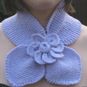May include: A light purple knitted scarf with a large flower design. The scarf is made of a soft, textured yarn and has a simple, elegant style.