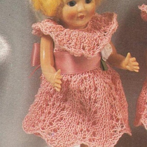 May include: A pink crocheted dress with a white lace trim and a pink satin sash. The dress is worn by a doll with blonde hair and blue eyes.