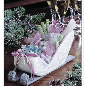 Op de afbeelding: Een witte gehaakte slee gevuld met snoep en verpakt in roze tule. De slee is versierd met zilveren glitter en een roze lint. De slee staat op een houten tafel met een groene krans en paarse kralen.