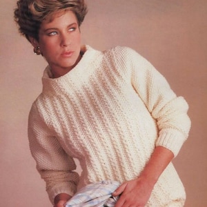 May include: A white knitted jumper with a high neck and a textured pattern. The jumper has long sleeves and is being worn by a person with short brown hair.