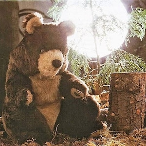 May include: A large brown teddy bear with a white face and black eyes sits in a woodland setting. The bear is sitting on a bed of brown leaves and twigs. There is a small tree stump behind the bear.