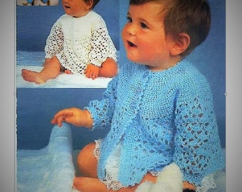 May include: A baby wearing a blue crocheted cardigan with a white crocheted undershirt. The baby is sitting on a white fluffy surface and holding a white stuffed animal.