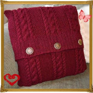 May include: A burgundy knitted pillow with a cable knit pattern and three decorative buttons. The pillow is on a brown surface.