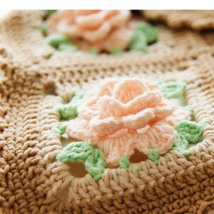 May include: A crocheted square with a light brown and beige background and a pink flower with green leaves in the center.