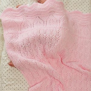 May include: A sleeping baby is nestled under a soft, light pink knitted blanket. The blanket features a delicate openwork pattern and a scalloped edge. The baby is wearing a white onesie, and the blanket is draped over a polka dot sheet.