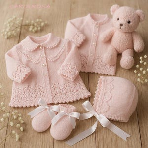 Op de afbeelding: Een collectie babykleding en accessoires in zachtroze. De set bevat twee gebreide vesten, een muts, babyslofjes met witte satijnen strikken en een teddybeer. De vesten hebben parelmoer knopen en kanten details.