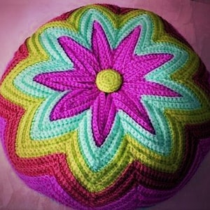 Op de afbeelding: Een gehaakt kussen in de vorm van een bloem met roze, groen en paars garen. De bloem heeft een knoop in het midden.