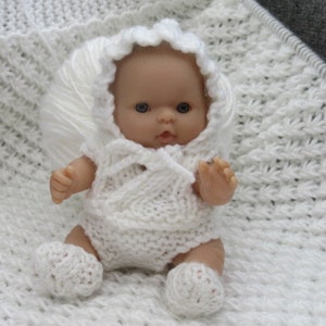 May include: A white knitted baby doll with a white knitted hat and booties. The doll is lying on a white knitted blanket.
