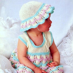 Op de afbeelding: Een baby draagt een gebreide jurk met een lichtblauw, roze en wit kleurpatroon. De jurk heeft een gegolfde zoom en een mouwloos ontwerp. De baby draagt ook een bijpassende gebreide muts met een gegolfde rand.