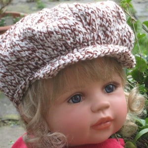 Può includere: Una bambola che indossa un cappello di lana marrone e bianco con una tesa. Il cappello ha un motivo testurizzato ed è fatto di filato.
