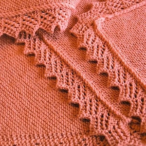 May include: Close-up of a coral-colored knitted blanket with a lacy border. The blanket is made with a soft, textured yarn and has a delicate, feminine feel.