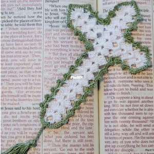 May include: A white crocheted cross bookmark with a sage green border and a matching sage green tassel at the bottom. The cross is placed on an open book with printed text. The bookmark features a delicate openwork pattern throughout the design.