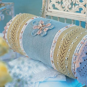 May include: A light blue knitted pillow with a crocheted flower design in shades of pink and blue. The pillow has a decorative trim of white and beige crocheted lace and beige buttons.