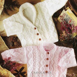 Puede incluir: Dos cárdigans de punto para bebé, uno blanco y otro rosa, con un patrón de punto de cable y botones de madera.