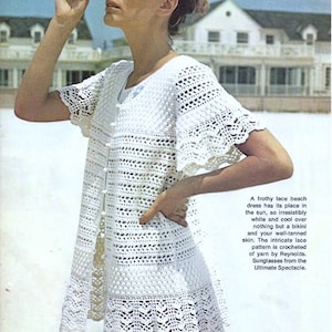 May include: A white crocheted beach dress with a delicate lace pattern. The dress is shown on a model wearing sunglasses and standing on a beach.