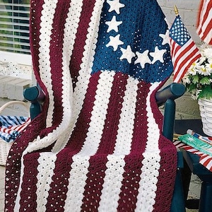 May include: A crocheted American flag blanket in red, white, and blue, draped over a wooden chair. The blanket features a traditional design with stars and stripes.