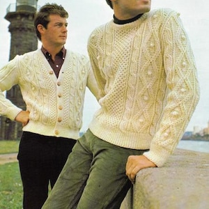 Op de afbeelding: Twee mannen dragen witte kabelgebreide truien. Een man draagt een cardigan met knopen en de andere draagt een trui met ronde hals. Beide mannen dragen een donkere broek.