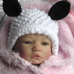 May include: A baby doll wearing a white crocheted hat with black felt ears. The hat has a textured, bobbly design. The doll is wearing a pink fuzzy jacket.