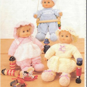 Könnte beinhalten: Drei gestrickte Puppenoutfits in Rosa, Blau und Gelb. Die Puppen tragen Hüte und haben bemalte Gesichter. Die Outfits sind alle in verschiedenen Stilen, eines ist ein Kleid, eines ein Overall und eines ein Pullover- und Hosenset. Die Puppen sitzen auf einer Holzoberfläche mit Holzspielzeug.