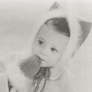 Puede incluir: Una fotografía en blanco y negro de un bebé con un gorro blanco de punto con capucha. El bebé mira hacia abajo y tiene una expresión seria en su rostro.