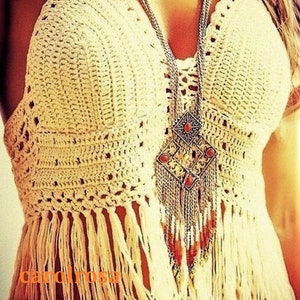 Puede incluir: Un top blanco de crochet con detalles de flecos y un collar de cadena plateada con un gran colgante adornado con piedras rojas. El top es de estilo halter y tiene un ajuste holgado.