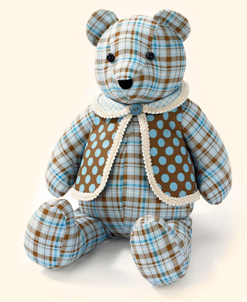 Pu&ograve; includere: Un orsacchiotto di peluche blu e marrone a quadri con un gilet marrone con pois blu e un bordo in pizzo bianco. L'orso &egrave; seduto con le gambe incrociate.