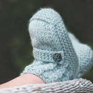 May include: A light blue knitted slipper with a decorative button closure. The slipper is made of a chunky knit yarn and has a folded-over cuff. The button is dark with a speckled design. The slipper is resting in a woven basket.