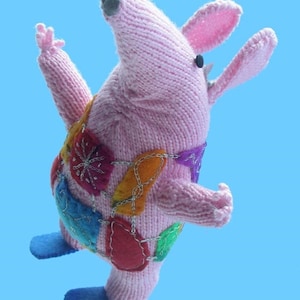 Puede incluir: Un juguete de peluche rosa tejido con un hocico largo y orejas grandes. El juguete lleva un chaleco de retazos colorido con varias formas y colores. El juguete tiene aletas de fieltro azul en los pies.
