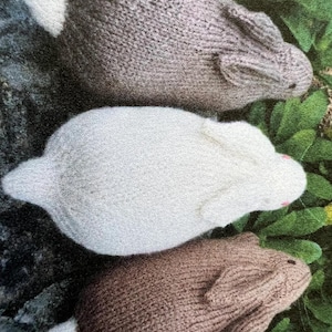May include: Three knitted bunny rabbits in different colors: gray, white, and brown. The bunnies are lying on a bed of green leaves.