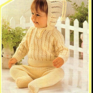 May include: A knitted baby outfit in a cream color. The outfit includes a long-sleeved sweater, pants, and a matching hat with a long tassel.